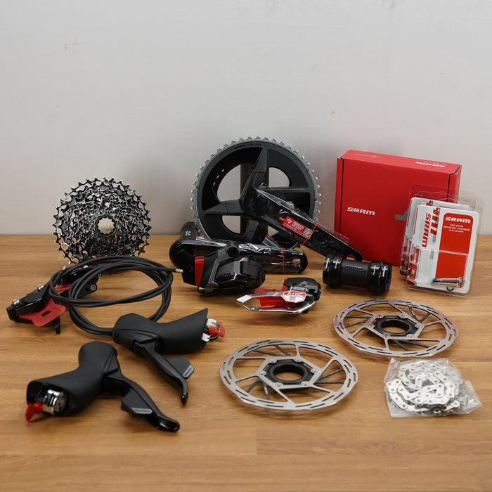 A laid-out bicycle component set on a wooden tabletop against a plain light wall, including a rear cassette, chain, brake lever, brake disc, brake rotor, headset, chainstay, headset cup, mounting hardware, and a red SRAMbox.