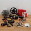 A bicycle component kit laid out on a wooden tabletop against a plain light wall, including a black crank arm, rear cassette, brake rotors, hand levers, a red SRAM box, and packaged parts.