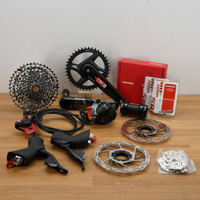 A bicycle component kit laid out on a wooden tabletop against a plain light wall, including a black crank arm, rear cassette, brake rotors, hand levers, a red SRAM box, and packaged parts.