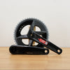 A black SRAM rival crankset with 2 chainrings sitting on a light brown wooden surface against a plain white background. 
