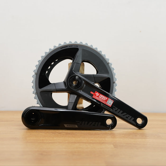 A black SRAM rival crankset with 2 chainrings sitting on a light brown wooden surface against a plain white background. 
