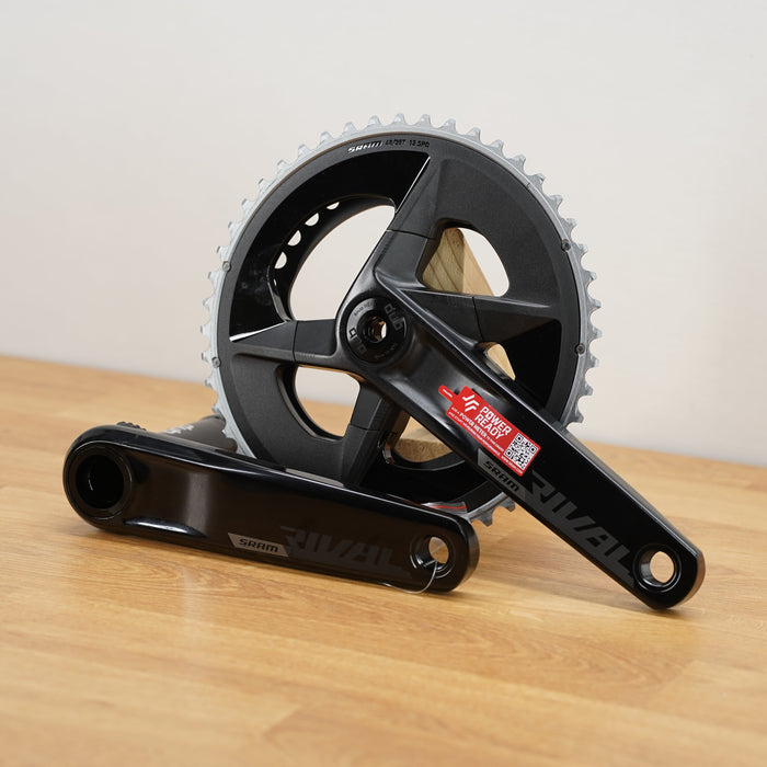 A black SRAM rival crankset with 2 chainrings sitting on a light brown wooden surface against a plain white background. 
