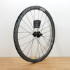 A carbon fiber road bicycle wheel with a deep section rim, thin spokes, and a central hub, standing upright on a light wood floor against a white wall.