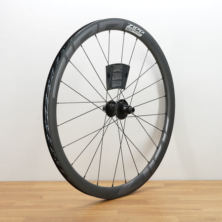 A carbon fiber road bicycle wheel with a deep section rim, thin spokes, and a central hub, standing upright on a light wood floor against a white wall.