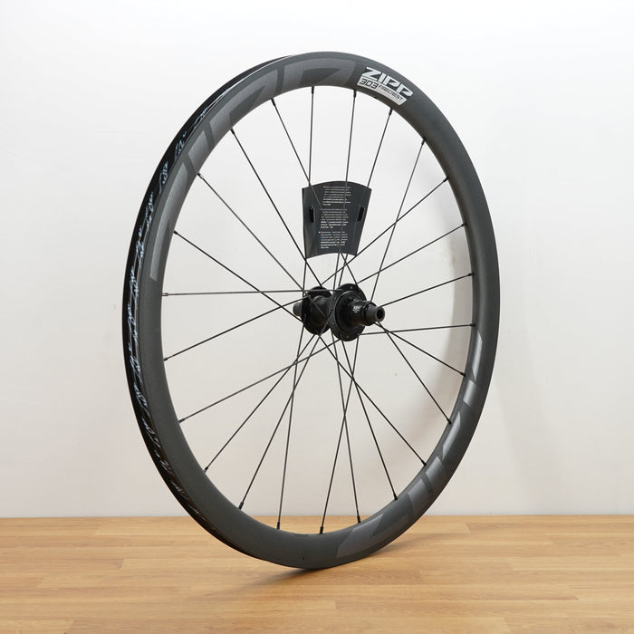 A carbon fiber road bicycle wheel with a deep section rim, thin spokes, and a central hub, standing upright on a light wood floor against a white wall.