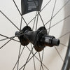 Close-up of a black bicycle hub and rim with thin black spokes against a light off-white background. The hub has visible branding including 'XDR'.