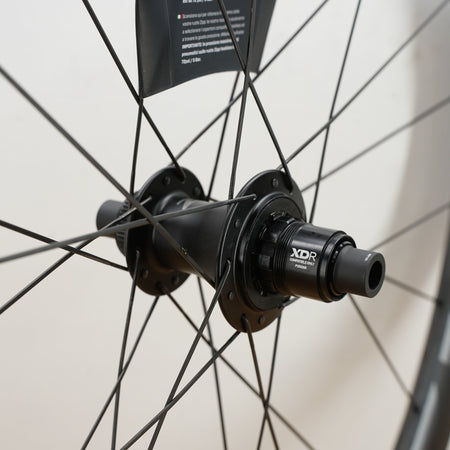 Close-up of a black bicycle hub and rim with thin black spokes against a light off-white background. The hub has visible branding including 'XDR'.