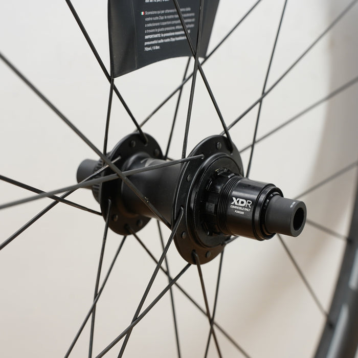 Close-up of a black bicycle hub and rim with thin black spokes against a light off-white background. The hub has visible branding including 'XDR'.
