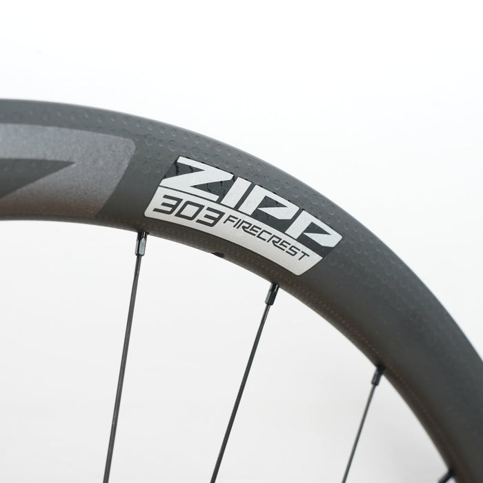 Close-up of a dark gray carbon fiber bicycle wheel rim with the white and gray ZIPP 303 Firecrest branding, set against a plain white background.
