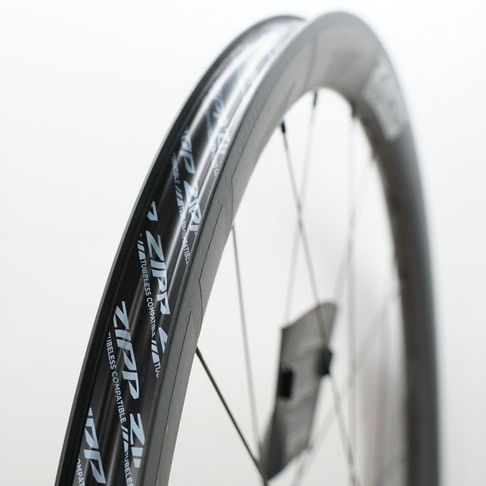 A close-up shot of a carbon fiber bicycle wheel rim and spokes, showing the brand name 'ZIPP' printed in white on the black rim, set against a clean white background.
