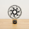 A black bicycle disc brake rotor mounted on a small wooden display block, set against a plain off-white wall and light wood floor.