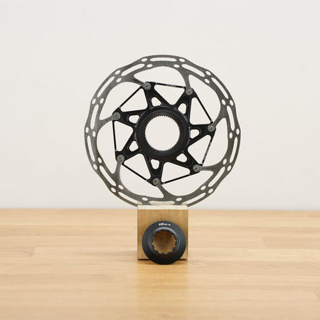 A black bicycle disc brake rotor mounted on a small wooden display block, set against a plain off-white wall and light wood floor.