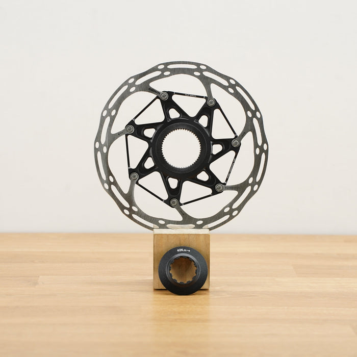 A black bicycle disc brake rotor mounted on a small wooden display block, set against a plain off-white wall and light wood floor.