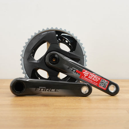 A black SRAM Force crankset with a red and white sticker, resting on a light wood surface against a white wall.