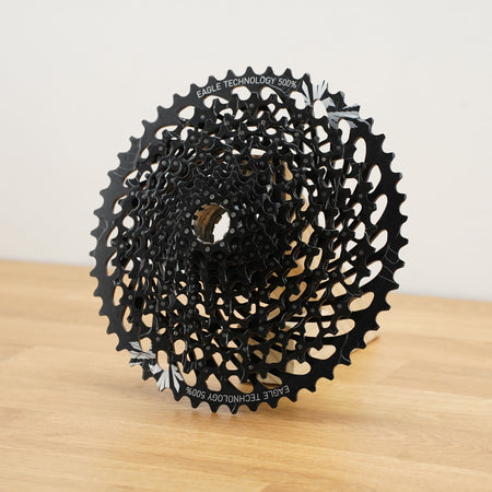 A black SRAM cassette photographed on a light wooden surface against a plain white wall. 