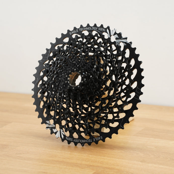 A black SRAM cassette photographed on a light wooden surface against a plain white wall. 