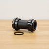 A black SRAM bottom bracket photographed on a light brown wooden surface against a plain white wall. 