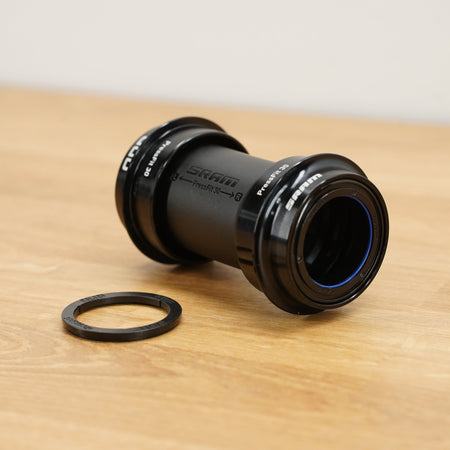 A black SRAM bottom bracket photographed on a light brown wooden surface against a plain white wall. 