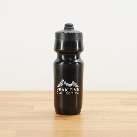 A black Peak Five Collective branded water bottle photographed on a light brown wooden surface against a plain white wall.