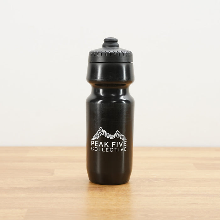 A black Peak Five Collective branded water bottle photographed on a light brown wooden surface against a plain white wall.