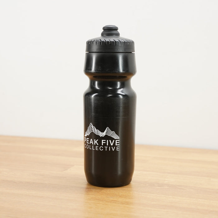 A black Peak Five Collective branded water bottle photographed on a light brown wooden surface against a plain white wall.