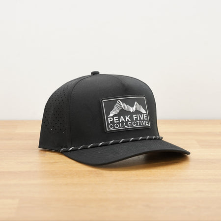 A black Peak Five Collective branded baseball cap photographed on a light brown wooden surface against a plain white wall .