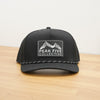 A black Peak Five Collective branded baseball cap photographed on a light brown wooden surface against a plain white wall .
