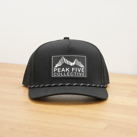 A black Peak Five Collective branded baseball cap photographed on a light brown wooden surface against a plain white wall .