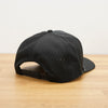 A black Peak Five Collective branded baseball cap photographed on a light brown wooden surface against a plain white wall .