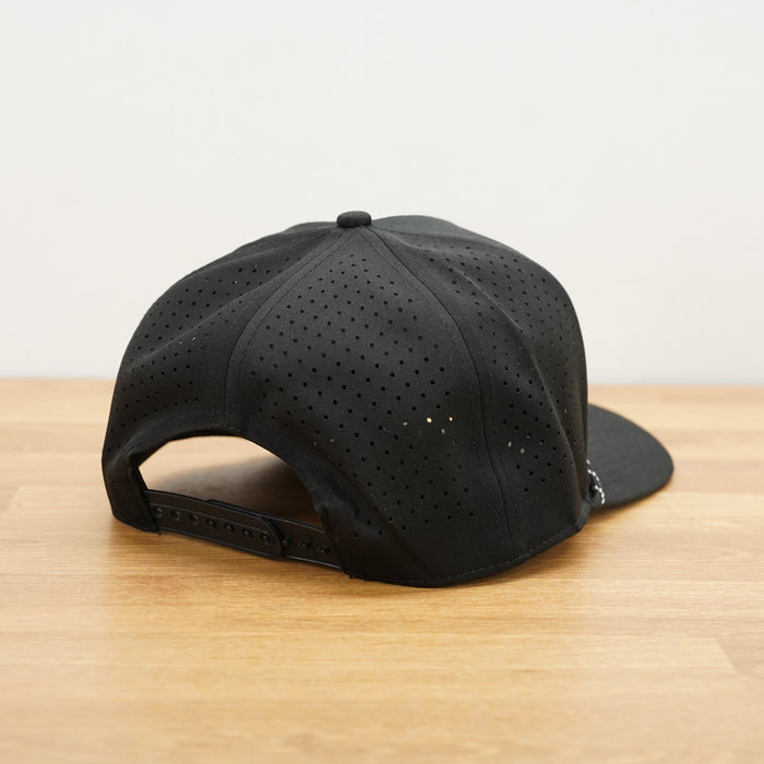 A black Peak Five Collective branded baseball cap photographed on a light brown wooden surface against a plain white wall .