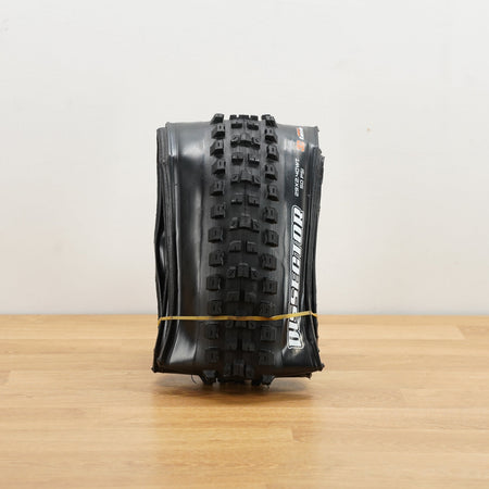 A black Maxxis Dissector tire photographed on a light brown wooden surface against a plain white wall. 