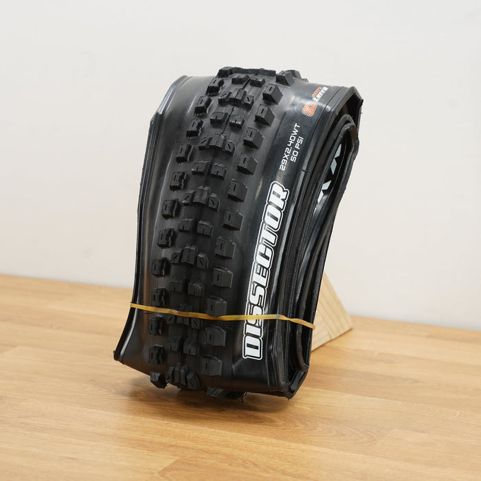 A black Maxxis Dissector tire photographed on a light brown wooden surface against a plain white wall. 