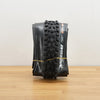 A black Maxxis Assegai mountain bike tire photographed on a light brown wooden surface against a plain white wall. 