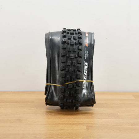 A black Maxxis Assegai mountain bike tire photographed on a light brown wooden surface against a plain white wall. 