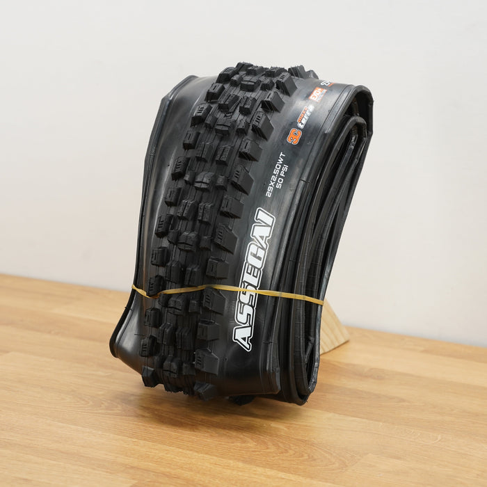 A black Maxxis Assegai mountain bike tire photographed on a light brown wooden surface against a plain white wall. 