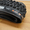 A black Maxxis Assegai mountain bike tire photographed on a light brown wooden surface against a plain white wall. 