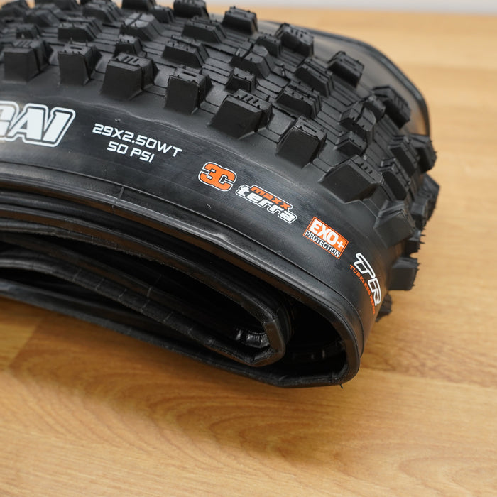 A black Maxxis Assegai mountain bike tire photographed on a light brown wooden surface against a plain white wall. 