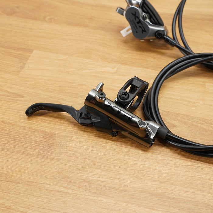 A bicycle brake lever assembly with a black cable resting on a light brown wooden floor, with a second brake lever visible in the background.
