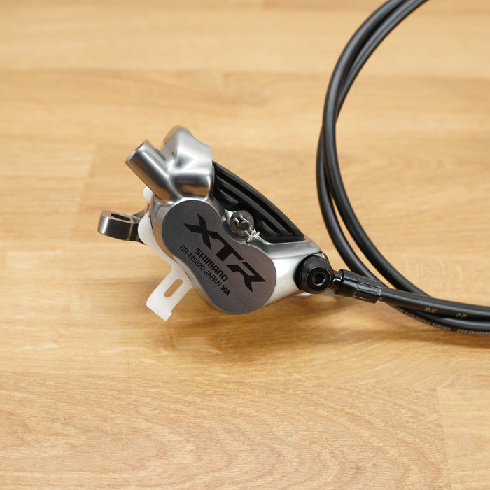 A close-up of a silver and black Shimano XTR hydraulic disc brake caliper with black brake lines, resting on a light brown wooden surface.