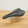 A black bicycle saddle with white details, displayed on a small wooden block stand on a light wood tabletop against a neutral wall.
