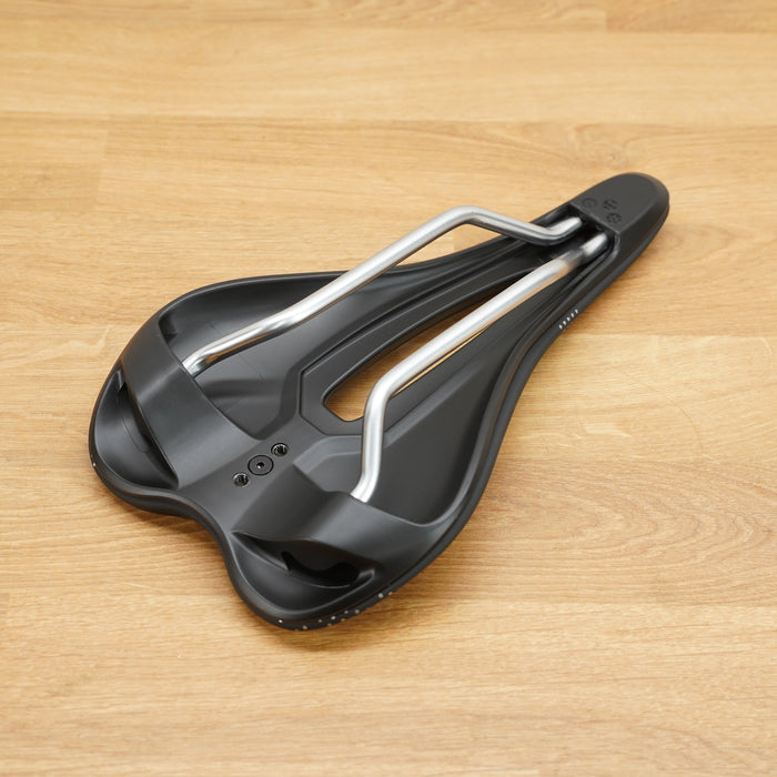 A black bicycle saddle with silver metal rails, photographed from a top-down perspective on a light brown wooden floor.