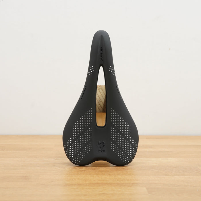 A black bicycle saddle shown upright on a light wood floor against a plain off-white wall.