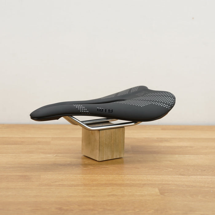 A black WTB bicycle saddle with white details, displayed on a small wooden block stand on a light wood tabletop against a neutral wall.