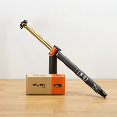 A black and gold Fox Transfer seat post  displayed diagonally on a black stand next to its cardboard accessories box on a light wood surface.