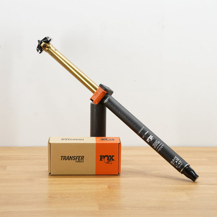 A black and gold Fox Transfer seat post  displayed diagonally on a black stand next to its cardboard accessories box on a light wood surface.