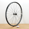 A black DT Swiss aluminum mountain bike wheel photographed on a light brown wooden surface against a plain white wall.