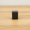 A small black rectangular Shimano Di2 battery photographed on a light brown wooden surface against a plain white wall.