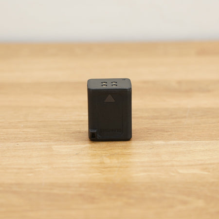 A small black rectangular Shimano Di2 battery photographed on a light brown wooden surface against a plain white wall.