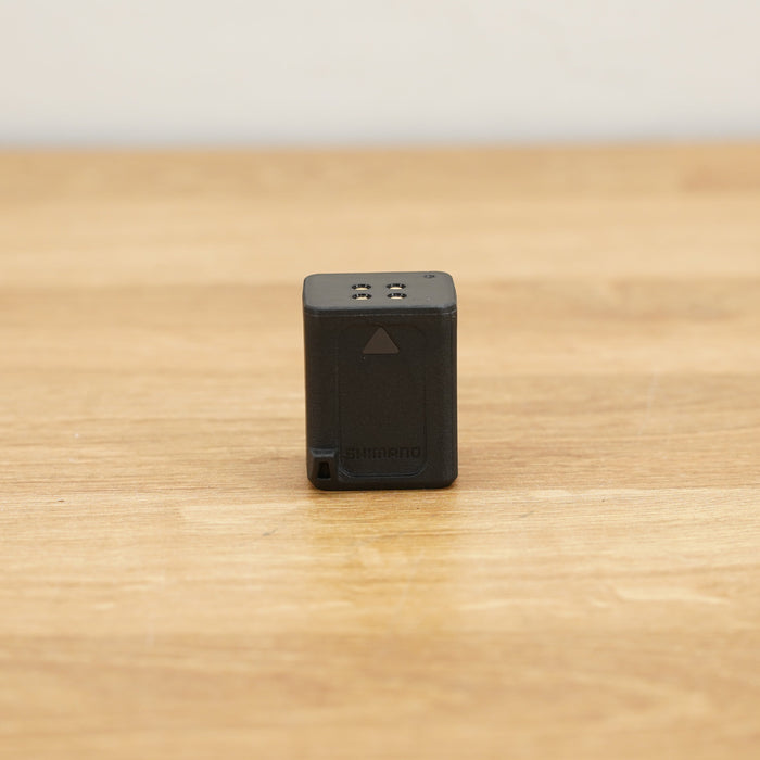 A small black rectangular Shimano Di2 battery photographed on a light brown wooden surface against a plain white wall.