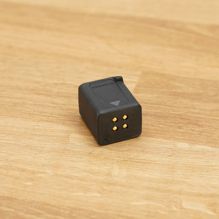 A small black rectangular Shimano Di2 battery photographed on a light brown wooden surface against a plain white wall.